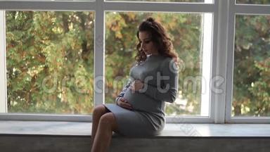 一位孕妇坐在家里靠窗的窗台上。她穿着漂亮的孕妇装